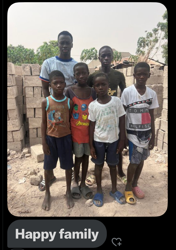 Six Gambian siblings stand side by side in front of a wall of bricks, barefoot and smiling softly, united in resilience and love.